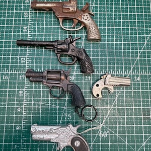 May include: A collection of five miniature metal toy pistols and revolvers in various finishes including rusted brown, matte black, and silver. The items are arranged on a green cutting mat with white grid lines and numerical markings.
