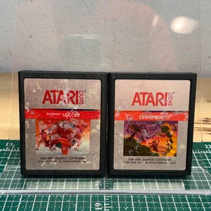 May include: Two black Atari 2600 video game cartridges. The left cartridge is labeled RealSports Soccer with a red graphic. The right cartridge is labeled Centipede with a multi-colored graphic. Both feature silver labels with wear.