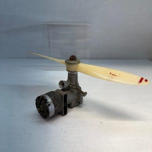 May include: A metallic model airplane engine featuring a ribbed cylinder head and a cream-colored plastic propeller attached to the front. The propeller blade displays the red text NYL and red markings near the tip, with a central metal nut.