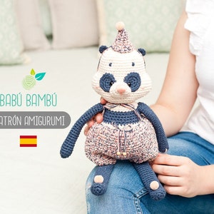 May include: A crocheted panda amigurumi toy wearing a pink and white hat and a blue and pink dress. The toy is being held by a person wearing blue jeans.