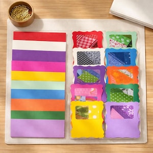 May include: A craft project layout featuring colorful paper strips and decorative frames. The paper strips are arranged in a vertical stack, showcasing a spectrum of colors. The frames, with scalloped edges, display patterned fabric samples. A small wooden bowl with gold wire and a white notepad are also present.