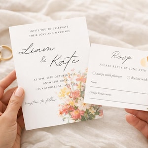 Op de afbeelding: Trouwkaartenset met bloemaccenten, met de namen "Liam & Kate". De uitnodiging bevat evenementdetails en een RSVP-kaart. Twee gouden ringen zijn zichtbaar. De set wordt door handen vastgehouden.