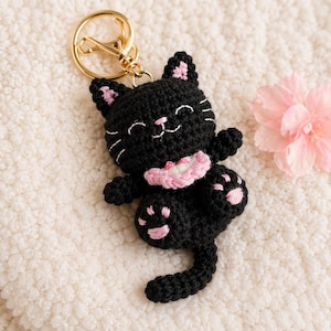 Peut inclure: Un porte-clés chat noir au crochet avec des détails roses sur les oreilles, les pattes et le collier. Le chat a des moustaches cousues blanches et un anneau de porte-clés doré.