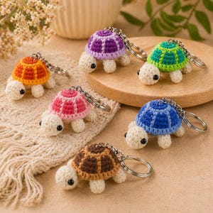 Può includere: Una collezione di sei portachiavi a forma di tartaruga fatti a mano all'uncinetto. Ogni tartaruga ha un guscio di colore diverso: arancione, viola, verde, blu, rosa e marrone. Hanno un corpo bianco, occhi neri e un anello portachiavi argentato. Esposti su legno e tessuto a maglia color crema.
