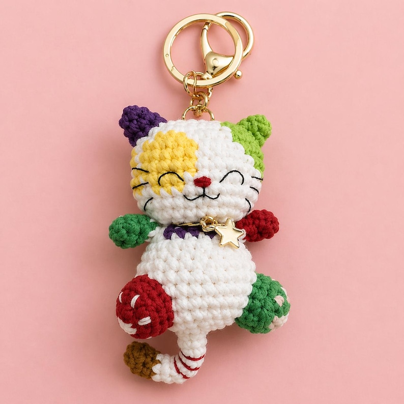 Pode incluir: Um chaveiro de gato de croch&ecirc; feito &agrave; m&atilde;o com um chaveiro dourado. O gato tem um corpo branco com detalhes coloridos em roxo, amarelo, verde e vermelho. Possui um pingente de estrela dourada, em um fundo rosa.