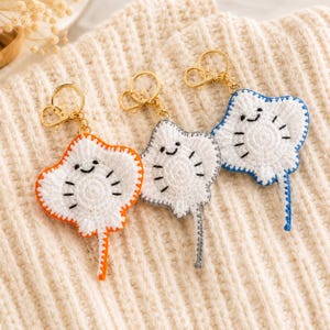 Op de afbeelding: Drie gehaakte pijlstaartrog-sleutelhangers met witte lichamen en zwart geborduurde ogen en kieuwen. Elke rog heeft een unieke randkleur: oranje, grijs of blauw. Elke sleutelhanger is voorzien van een goudkleurige metalen karabijnhaak.