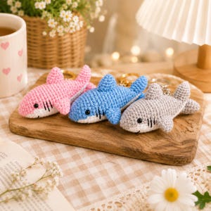 May include: Three handmade crochet shark keychains in pink, blue, and gray. Each shark features a white underbelly, black embroidered eyes, and three small black gill slits on the side. A gold-toned metal keyring is attached to the top of each.