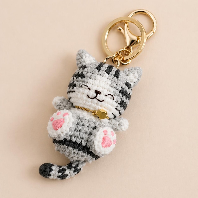 Pode incluir: Chaveiro de gato de croch&ecirc; em cinza e branco com listras pretas. O gato tem almofadas de pata rosa e uma coleira dourada. O chaveiro tem um fecho e um porta-chaves dourados.