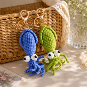 May include: Two crochet squid keychains, one blue and one green, each featuring large white eyes with black pupils and a gold-toned metal lobster clasp attached to a circular ring, crafted with detailed yarn stitching and multiple dangling tentacles.