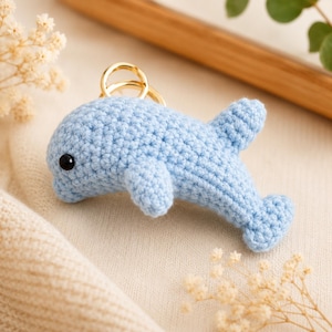 Könnte beinhalten: Ein hellblauer, gehäkelter Delfin-Schlüsselanhänger mit einem kleinen, runden schwarzen Auge und einem goldfarbenen Metallring an der Oberseite. Der Delfin ist in einer strukturierten Maschenoptik gefertigt und zeigt Rücken- und Seitenflossen.