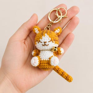 Pode incluir: Um chaveiro de gato de croch&ecirc; feito &agrave; m&atilde;o, em tons de laranja, branco e castanho. O gato tem uma coleira com sino dourado e um anel de chaveiro dourado. O gato tem um rosto sorridente e orelhas pequenas e pontudas. O chaveiro &eacute; um pequeno item feito &agrave; m&atilde;o.