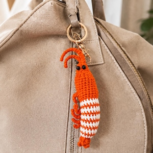 May include: A crocheted shrimp keyring featuring a vibrant orange head and body with alternating white and orange striped segments along the tail. The shrimp has two long antennae and small black bead eyes, attached to a gold-toned metal ring.