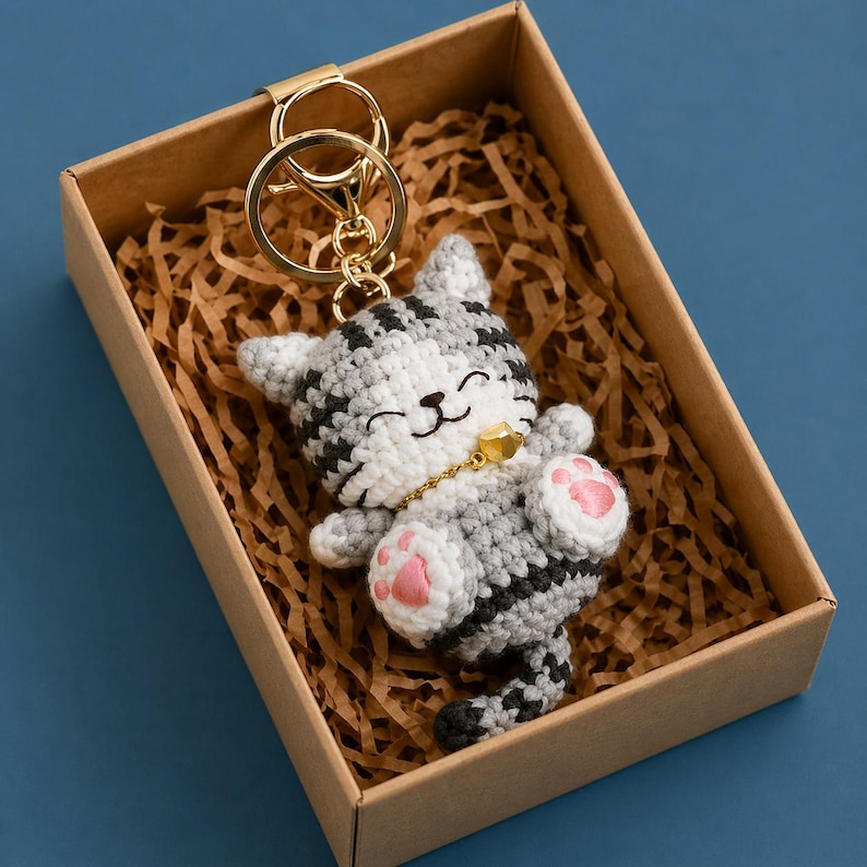 Pode incluir: Chaveiro de gato de croch&ecirc; em uma caixa de presente. O gato &eacute; cinza, branco e preto com almofadas de pata rosa e uma coleira dourada. O chaveiro tem um anel e um fecho dourados. A caixa de presente &eacute; marrom e cheia de papel picado.