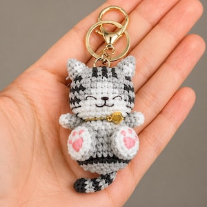 Pode incluir: Chaveiro de gato de croch&ecirc; em cinza, branco e preto com almofadas de pata rosa e uma campainha dourada. O gato tem um rosto sorridente e est&aacute; preso a um chaveiro dourado. Um acess&oacute;rio artesanal e caprichoso.