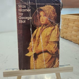 Puede incluir: Un libro de bolsillo vintage titulado "Silas Marner" de George Eliot. La portada presenta un retrato de un niño con gorro y vestido. El libro se exhibe en un pequeño caballete de madera.