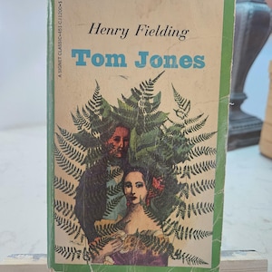 Puede incluir: Libro de bolsillo vintage titulado "Tom Jones" de Henry Fielding. La portada presenta ilustraciones de dos personas rodeadas de helechos. El libro tiene un borde verde y el texto "A SIGNET CLASSIC" es visible.