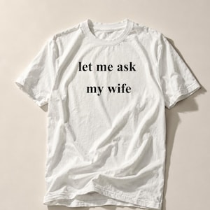 Może przedstawiać: Biała koszulka z czarnym napisem "let me ask my wife". Koszulka ma krótkie rękawy i okrągły dekolt. Materiał jest lekko pognieciony.