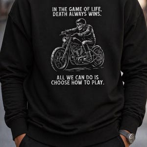 May include: Black sweatshirt with a white graphic of a skeleton on a motorbike. The text reads "IN THE GAME OF LIFE, DEATH ALWAYS WINS. ALL WE CAN DO IS CHOOSE HOW TO PLAY" above and below the illustration.