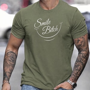 May include: Olive green crew neck t-shirt with the text "Smile Bitch" in white script. The shirt is worn with blue jeans. The person wearing the shirt has tattoos on both arms.