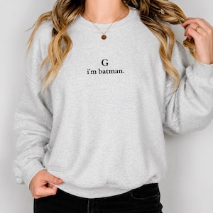 May include: A light gray sweatshirt with the text "G i'm batman." in black. The sweatshirt is a crew neck style and appears to be made of a soft material. The letter "G" is above the text.