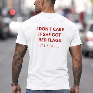 以下が含まれることがあります： 赤い文字で「I DON'T CARE IF SHE GOT RED FLAGS I'M A BULL」と書かれた白いTシャツ。Tシャツは人が着ています。その人はブルージーンズを履いており、腕にタトゥーがあります。