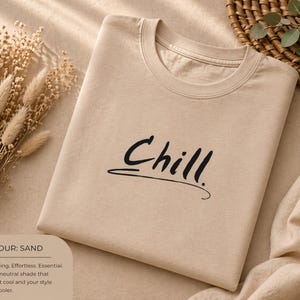 Peut inclure: Un t-shirt couleur sable avec le mot "Chill" en écriture noire. Le t-shirt est plié proprement et est entouré de fleurs séchées et d'un panier en osier. La couleur est décrite comme décontractée et sans effort.