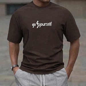 Pode incluir: Camiseta marrom de manga curta com a escrita branca "go yourself". A camiseta tem gola redonda. O modelo usa calças de moletom cinza e um relógio. O estilo é casual.
