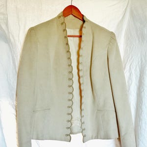 May include: A light beige jacket with a scalloped trim and decorative buttons. The jacket has a mandarin collar and two front pockets. It is hanging on a wooden hanger against a white background.