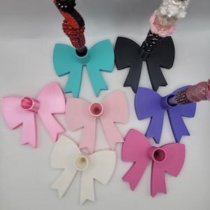 May include: A collection of colorful bow-shaped stands in various colors, including teal, black, pink, purple, and white. Each stand has a cylindrical holder. The stands are arranged on a white surface.