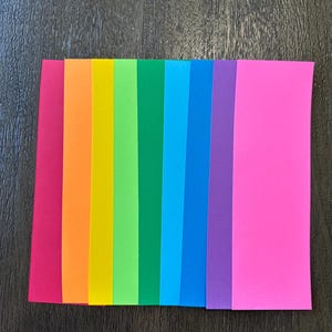 May include: A collection of nine rectangular paper strips arranged side by side in a spectrum of colors including magenta, orange, yellow, lime green, forest green, cyan, blue, purple, and hot pink, creating a vibrant rainbow-like sequence.