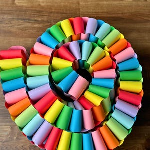 May include: A colorful, spiral-shaped paper craft. The artwork features a vibrant array of rainbow colors, including red, orange, yellow, green, blue, and purple. The paper is rolled into cone shapes, creating a textured, layered effect.