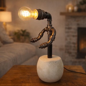 Puede incluir: Una lámpara de mesa de estilo industrial único. La lámpara presenta una bombilla en la parte superior, sostenida por tuberías y accesorios de metal negro, y una base de hormigón. La lámpara está sobre una mesa de madera, con un sofá y una chimenea al fondo.