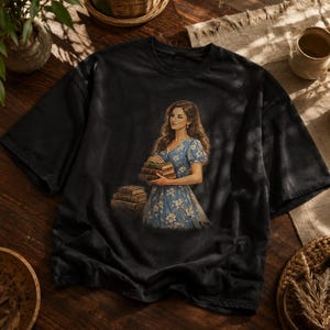 Peut inclure: Un t-shirt noir à manches courtes avec une impression graphique représentant une personne aux longs cheveux ondulés portant une robe bleue à fleurs. La personne tient une pile de trois livres, tandis qu'une autre pile de trois livres est posée au sol.
