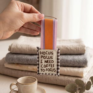 Pode incluir: Um marcador de livros decorativo com o texto "Hocus Pocus I Need Coffee To Focus". O marcador de livros tem um padrão xadrez marrom e branco, com fitas laranja, rosa e roxa. Uma pequena xícara e pires de café de cerâmica estão em primeiro plano.