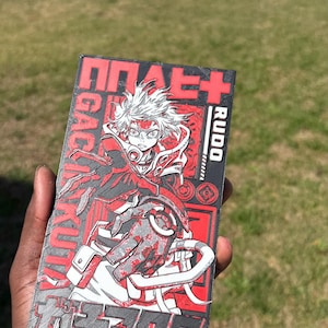 May include: A rectangular graphic in red, black, and grey, featuring an anime character with white hair and red details. Japanese text and the word "Rudo" surround the character. The artwork is held in a hand.