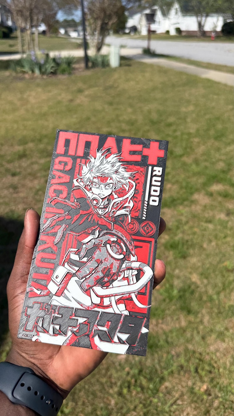 May include: A rectangular graphic in red, black, and grey, featuring an anime character with white hair and red details. Japanese text and the word "Rudo" surround the character. The artwork is held in a hand.