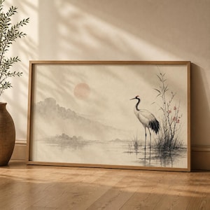 Op de afbeelding: Een rechthoekige ingelijste kunstdruk met een landschap in aquarel-stijl met een kraanvogel die in ondiep water staat. De scène bevat een bleke zon, mistige bergen en hoog riet met kleine roze bloesems, geplaatst in een lichte houten lijst.