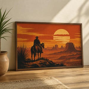 May include: A rectangular framed wall art print depicting a desert landscape at sunset. A rider on horseback wearing a wide-brimmed hat stands on a rocky ridge overlooking mesas and buttes under a large, glowing orange sun in the sky.