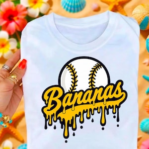May include: White t-shirt featuring a baseball graphic with the word "Bananas" in yellow dripping font. The baseball is white with black stitching, set against a black outline. The shirt is held by a person with multiple rings.