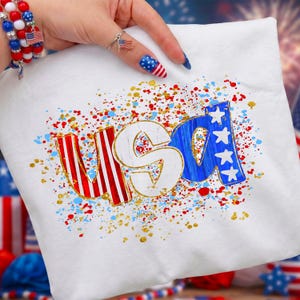 May include: White t-shirt with the word "USA" in red, white, and blue, with gold accents and a splatter design. The letters are stylized with the "U" and "A" in red and white stripes, and the "S" in white with a gold outline. The "A" has three white stars.