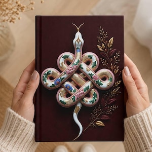 May include: A dark maroon hardback book featuring a central illustration of a white snake coiled into an intricate knot pattern. The snake is decorated with colourful floral motifs in pink, blue, and green, accompanied by a golden leafy branch design.