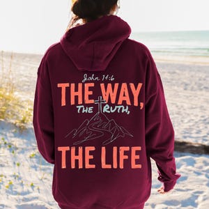 以下が含まれることがあります： 背面にJohn 14:6、THE WAY、THE TRUTH、THE LIFEというオレンジと水色の文字がプリントされたマルーンカラーのパーカー。デザインには、山の小道と頂上の十字架の線画が含まれています。