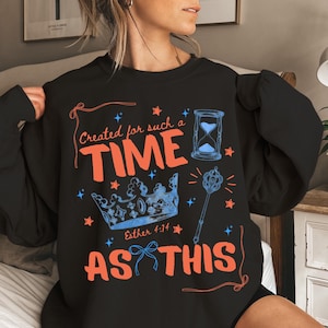 以下が含まれることがあります： 「Created for such a TIME AS THIS」および「Esther 4:14」というテキストがプリントされた黒いクルーネックのスウェットシャツ。デザインには、青い王冠、青い砂時計、青い笏、そして小さなオレンジ色と青色の星が含まれています。