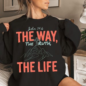 以下が含まれることがあります： コーラルとライトブルーで「John 14:6, THE WAY, THE TRUTH, THE LIFE」というテキストがプリントされた黒の長袖スウェットシャツ。前面中央には、頂上に十字架が描かれた山の線画グラフィックがデザインされています。