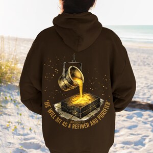 May include: A dark brown hooded sweatshirt featuring a graphic on the back of a metal bucket pouring molten gold into a mold shaped like a cross. The text reads, "HE WILL SIT AS A REFINER AND PURIFIER" and "MALACHI 3:3" in a circular gold font.