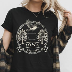 Może przedstawiać: Czarny t-shirt z krótkim rękawem z białym nadrukiem graficznym. Wzór przedstawia ptaka w locie, dwa dęby, zarys stanu Iowa oraz wstęgę z napisem IOWA EST. 1846 umieszczoną wewnątrz okrągłej obwódki przypominającej linę.