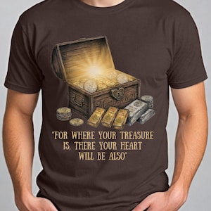 May include: Brown t-shirt featuring an illustration of an open treasure chest overflowing with coins and gold bars. The text "FOR WHERE YOUR TREASURE IS, THERE YOUR HEART WILL BE ALSO" is printed below the chest. The shirt has a crew neck.