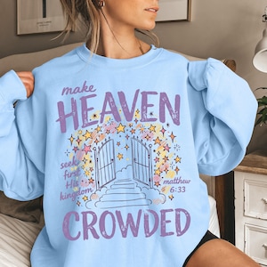 以下が含まれることがあります： 「make HEAVEN CROWDED」および「seek first His kingdom matthew 6:33」というテキストがプリントされた、ライトブルーのクルーネックトレーナー。開いた門、階段、カラフルな星のイラストが描かれています。