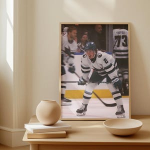 May include: Framed photograph of a hockey player in a white and green uniform, number 2, on the ice. The player is in a skating stance, holding a hockey stick. The background shows other players and spectators. The frame is light brown.