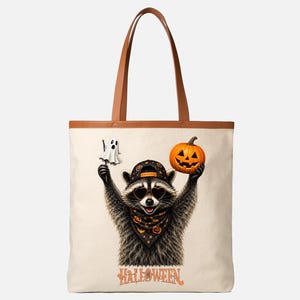 May include: A cream-colored tote bag with brown handles featuring a graphic of a raccoon wearing a baseball cap, heart-shaped sunglasses, and a patterned bandana. The raccoon holds a ghost and a jack-o'-lantern, with the word HALLOWEEN below.